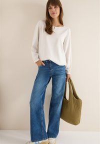 Hvid langærmet bluse med rund halsudskæring, parret med vide blå denimjeans og en grøn skuldertaske med fyldige skuldre. Sneakers med detaljer.