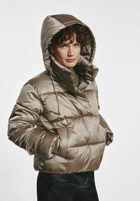 Peleryna z puchowym wypełnieniem w błyszczącym oliwkowym kolorze, z kapturem, pikowanym wzorem i elastycznymi mankietami. Widoczne są dwie boczne kieszenie.