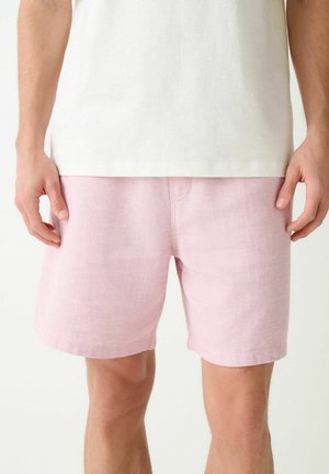 Mann trägt hellrosa texturierte Shorts und ein schlichtes weißes Hemd, steht mit locker an den Seiten hängenden Händen.
