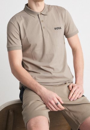 Mężczyzna w beżowej koszulce polo z logo "BOSS" i pasujących beżowych szortach siedzi na drewnianej ławce, ręka oparta na nodze.