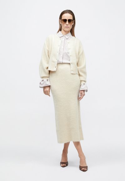 Moteris, vilkinti pereaugintą smėlio spalvos megztinį, dryžuotą marškinėlį, derančią smėlio spalvos midi sijoną, rudus atvirus kulnius ir akinius nuo saulės, stovinti prieš baltą foną.