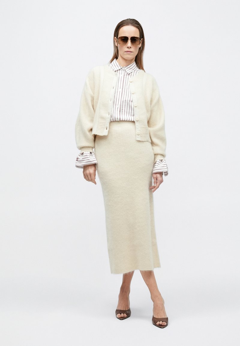 Woman wearing oversized beige cardigan, striped shirt, matching beige midi skirt, brown open-toe heels, and sunglasses standing against white background.