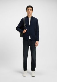 Granatowa kurtka zapinana na zamek, biała koszula, ciemne dżinsy i białe sneakersy. Model trzyma torbę, stoi na jednolitym tle.