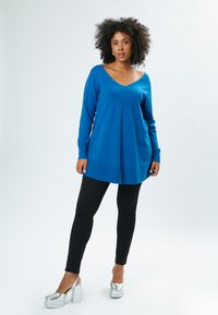 Blauer Pullover mit langen Ärmeln, V-Ausschnitt und strukturierten Details. Kombiniert mit schwarzen Leggings und silbernen Plateauschuhen.