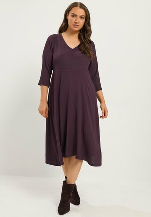 Femme portant une robe prune longueur genou avec manches trois-quarts et des bottines noires, debout devant un fond uni.