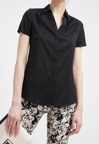 Camicia nera a maniche corte con bottoni frontali e colletto, abbinata a pantaloni con motivo floreale. La camicia ha una texture liscia.