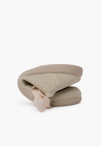 Zapatillas beiges con una textura tejida, parte superior de tela suave y suela de goma flexible. Presentan un diseño plegado con un acento de tela.