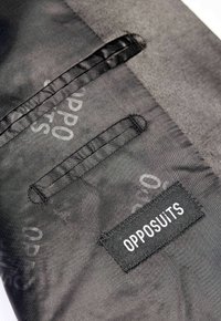 Tessuto nero con texture liscia, caratterizzato da una tasca verticale e un'etichetta con la scritta "OPPOSUITS" in bianco. Il design è minimalista ed elegante.