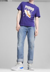 Person in einem lila Puma-T-Shirt mit Schuhmotiv, hellblauen, hochgekrempelten Jeans und weißen Sneakers mit blauen Akzenten, der vor einem schlichten Hintergrund steht.