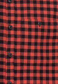 Chemise à carreaux rouge et noir en tissu tissé, avec de grands motifs à carreaux, six boutons noirs et une poche poitrine.