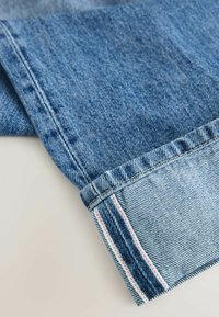 Jambe de jean en denim bleu retroussée pour montrer le bord selvedge avec des coutures blanches et rouges sur un fond clair.