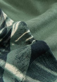 Zbliżenie zielonej tkaniny o miękkiej, dzianinowej fakturze z nadrukiem przedstawiającym ciemne i jasne abstrakcyjne wzory.