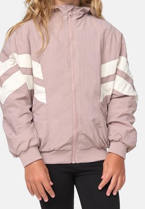 Kind draagt licht mauve ritsjack met witte chevronstrepen op de mouwen, gecombineerd met een zwarte broek, staand tegen een effen achtergrond.