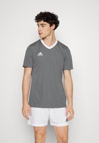 Camisa atlética gris de manga corta, con escote en V y cuello y detalles blancos. Presenta el logo de Adidas en el lado izquierdo del pecho.