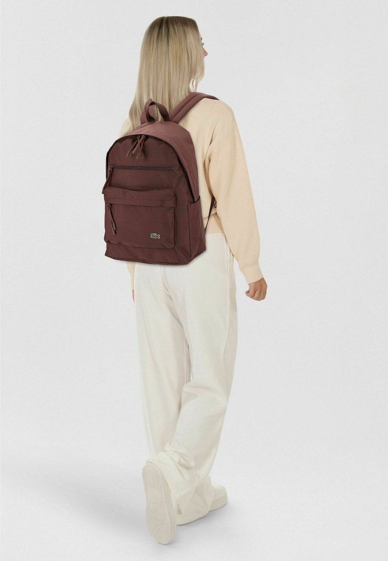 Maroon backpack with a textured fabric, featuring a front pocket and zipper closure. Adjustable straps and a small logo visible on the front.