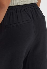 Schwarze Leinenhose mit strukturierter Oberfläche, elastischem Bund und zwei Gesäßtaschen sowie einem dezenten Nahtdetail an den Seiten.