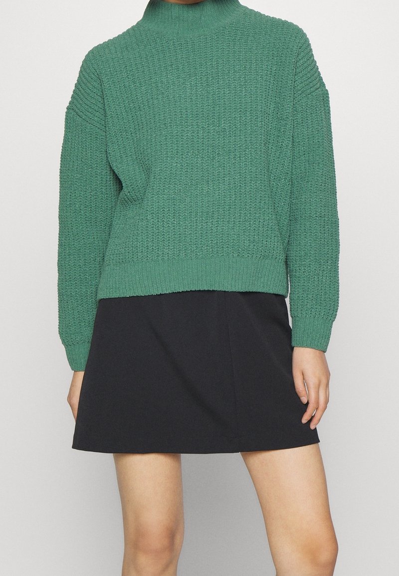 Personne portant un pull vert texturé à manches longues, rentré dans une jupe patineuse noire, avec les jambes nues visibles en dessous.