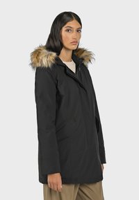 Svart parkas med huva som är fodrad med fuskpäls, dragkedja och knappstängning, två framfickor och en figursydd passform. Slät, vattenavvisande tyg.