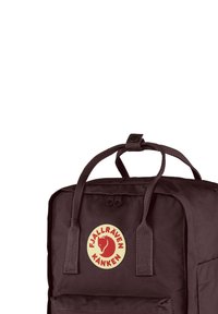 Dunkelvioletter Kånken-Rucksack mit einem runden Logo, das ein rotes Fuchsdesign zeigt, zwei stabilen oberen Griffen und einer Frontreißverschlusstasche. Strukturierter Stoff.