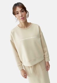 Light beige sweatshirt featuring a round neck, long sleeves, and a textured graphic print across the chest. Soft, smooth fabric.