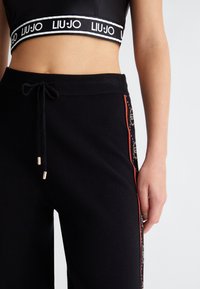Schwarze Jogginghosen mit rotem Seitenstreifen und goldenen Hardware-Akzenten, ausgestattet mit einem Kordelzugbund. Das Material wirkt weich und hat eine glatte Textur.
