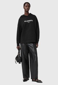 Felpa nera con tasca davanti e logo bianco, abbinata a pantaloni in pelle nera lucidi e scarpe nere. Tiene una borsa nera.