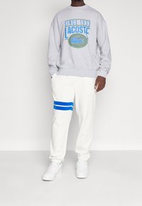 Felpa grigia con grafica blu "LACOSTE SINCE 1933", abbinata a pantaloni bianchi con strisce blu. Il modello indossa sneakers bianche.