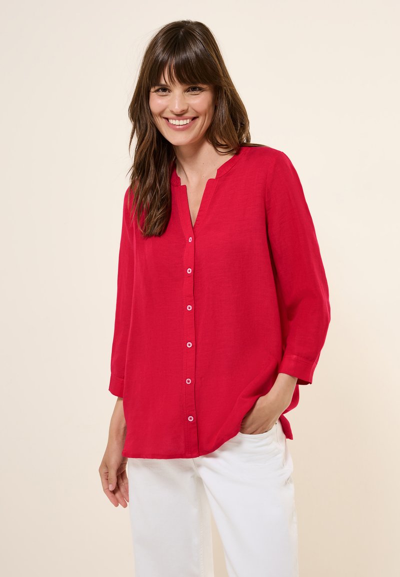 Camicia rossa con bottoni, scollo a V, maniche a tre quarti e vestibilità comoda, realizzata in tessuto leggero. Modello in piedi contro uno sfondo neutro.