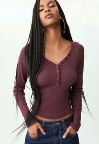 Burgundy long-sleeve ribbed top featuring a v-neckline, button detail, and a fitted design. Paired with dark blue denim jeans.