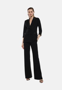 Blazer nero doppiopetto con scollo a V profondo, maniche a tre quarti, abbinato a pantaloni neri a gamba larga. Texture liscia, dettagli minimi.