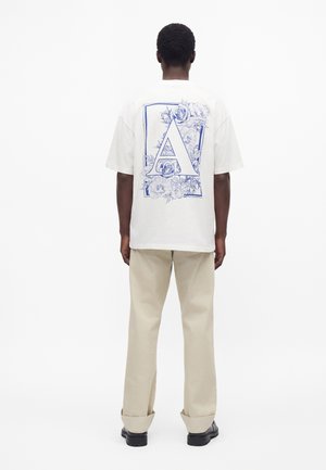 Mann trägt weißes T-Shirt mit großem blauem Buchstaben A und floralem Muster auf dem Rücken, beige Hose und schwarze Schuhe, steht mit dem Rücken zur Kamera.