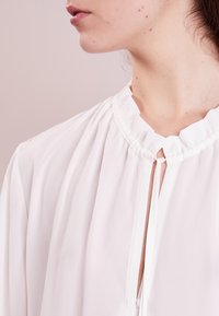 White blouse featuring a high neck with ruffled collar, vertical pleats, and a keyhole opening at the front. Soft, lightweight fabric.