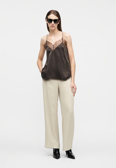 Γυναίκα που φορά μαύρο καλοκαιρινό τοπ με διάφανα περιθώρια, μπεζ παντελόνι φαρδύ πόδι, μαύρα μυτερά μποτάκια και μαύρα γυαλιά ηλίου σε απλό λευκό φόντο.