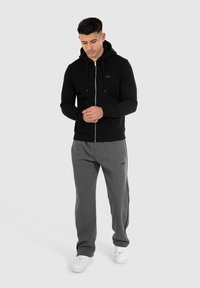 Sweat à capuche noir zippé avec une texture douce, associé à un pantalon de survêtement gris avec des rayures sur les côtés. Des baskets blanches complètent la tenue.