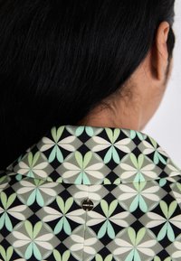 Hemdbluse mit einem geometrischen Blumenmuster in grün, weiß und schwarz. Enthält ein kleines, rundes Metalldetail mit Logo.