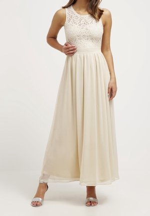 Dlouhé krémové šaty s krajkovým živůtkem, bez rukávů a s volným sukněm. Mají přiléhavý pas a jemný lesk. Styl doplňují stříbrné sandály.