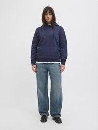 Felpa blu navy con coulisse e tasca frontale, indossata sopra una camicia bianca. Abbinata a jeans a gamba larga blu chiaro e scarpe nere.