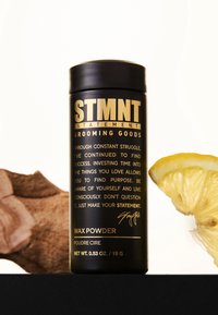 Svart cylindrisk behållare för vaxpulver, med guldfärgad text och logotyp. Inkluderar en citronskiva och ett texturerat brunt material bredvid.