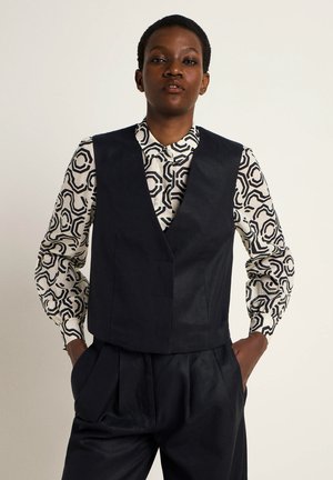 Marineblaues Leinenweste mit tailliertem Schnitt über einem gemusterten schwarz-weißen Hemd. Das Hemd hat ein geometrisches Design und einen Button-Down-Kragen.