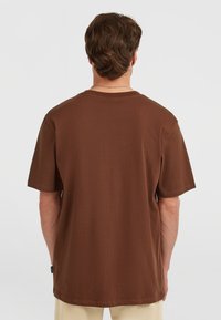 Brun bomulds t-shirt med korte ærmer, rund halsudskæring og lige kant. Har et sidelabel nær den venstre søm.