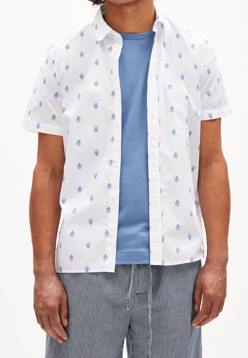 Camicia bianca a maniche corte con motivo floreale blu, con bottoni sul davanti, indossata sopra una maglietta a collo rotondo azzurra. Pantaloni a righe visibili sotto.