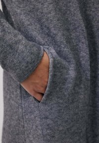 Main partiellement insérée dans la poche latérale d'un vêtement à manches longues en tricot gris doux, montrant la texture du tissu et les détails des coutures.