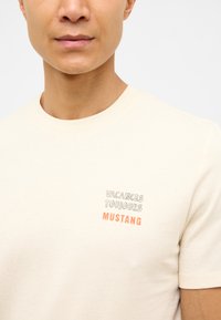 Beige kokvilnas T-krekls ar apaļu kakla izgriezumu. Izceļas ar gaišiem uzrakstiem "VACANCES TOUJOURS" un "MUSTANG" oranžā un pelēkā krāsā.