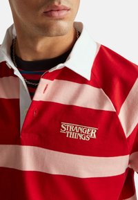 Mężczyzna w czerwono-różowej koszuli w paski z białym kołnierzykiem i wyhaftowanym logo "Stranger Things" na piersi.