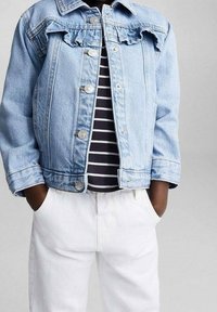 Ljusblå denimjacka med rynkad axel, silverknappar och två framfickor, bärs över en svart och vit randig skjorta och vita byxor.