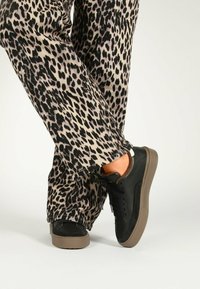 Sneakers neri con tomaia testurizzata e accenti marroni, abbinati a pantaloni a gamba larga con stampa leopardo. Le scarpe presentano una suola in gomma e lacci.
