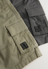 Deux paires de shorts cargo en olive et gris foncé, avec plusieurs poches et des étiquettes noires avec du texte, fabriquées en tissu lisse.