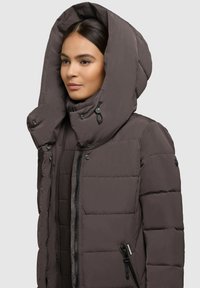 Donna che indossa una giacca invernale imbottita marrone scuro con un grande cappuccio e chiusura con zip, su uno sfondo chiaro e semplice.