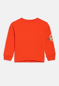 Svetlo oranžové tričko s dlhými rukávmi, okrúhlym výstrihom a okrúhlym pretekárskym logom na pravom rukáve.