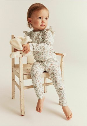 Blumenmuster Baumwoll-Pijama-Set mit Rüschendetails; heller Hintergrund, weiße Basis und sanftes Pastellmuster von Tieren und Blumen. Kind hält Stofftier.